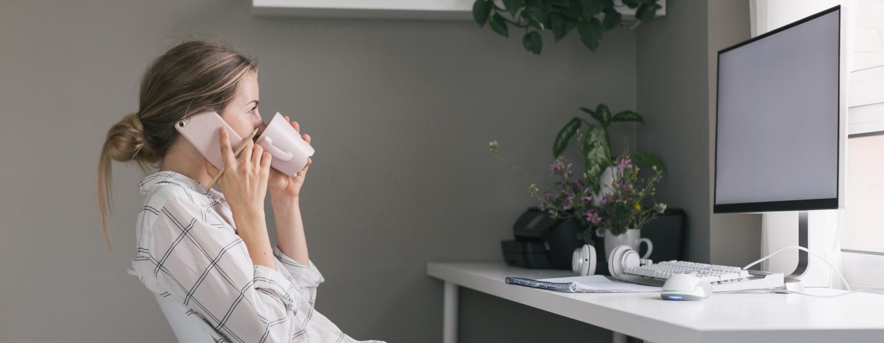 a woman sitting at a desk talking on a cell phone while drinking coffee